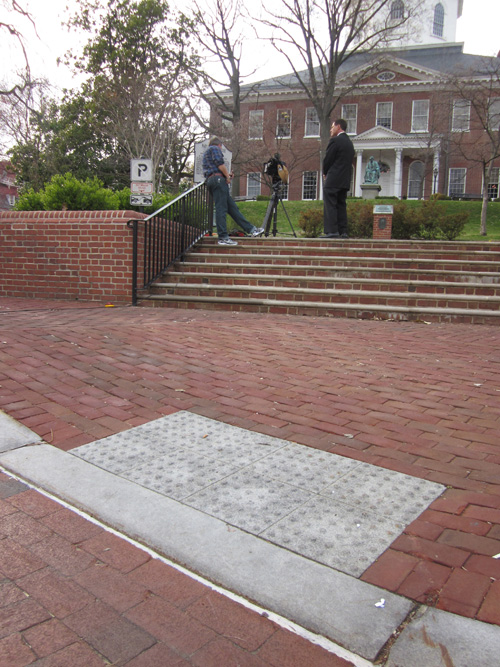 Capitol: Three photos show a sidewalk in front of a wide stairway with a retaining wall about 4 feet high on each side of the stairs.  The street, sidewalk, wall stairs, and ramp going around the stairs are all made of red brick.  The brick slopes down to the street to form a curbramp about 4 feet wide.  The detectable warning is made out of two rows of 4 white cement tiles which are each about a foot square with 64 truncated domes (6 on each side of the tile).